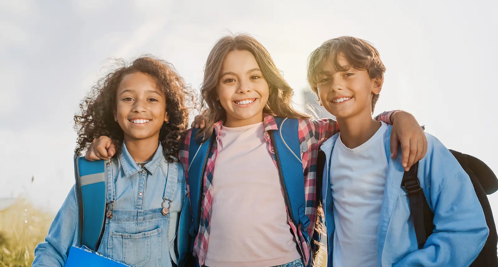 Three smiling children with backpacks in Magnolia TX, standing outdoors, arms around each other.
