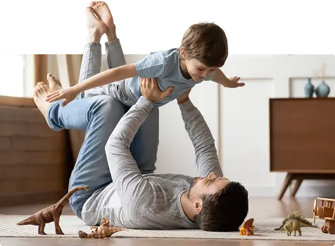 An adult lies on the floor in Magnolia TX, lifting a smiling child amid toy dinosaurs.